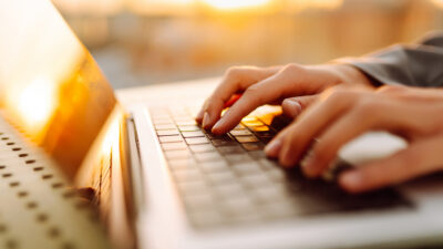 Close-up de mãos digitando em um laptop. Mãos de uma jovem trabalhando em um laptop sob os raios do pôr do sol. Conceito de trabalho freelancer e tecnologia.