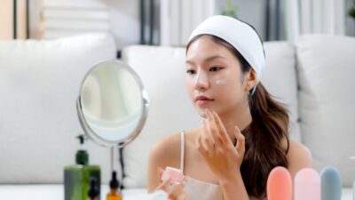 Jovem asiática aplicando creme no rosto para rugas enquanto olha espelho na sala de estar k-beauty cosméticos coreanos skincare