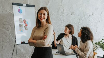 empreendedora sentada em uma mesa de reuniao enquanto duas profissionais apontam para uma apresentação no fundo