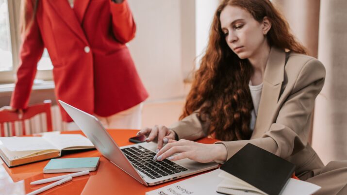 Duas mulheres em traje de negócios focadas no trabalho em um laptop, dentro de casa, em uma mesa redonda.