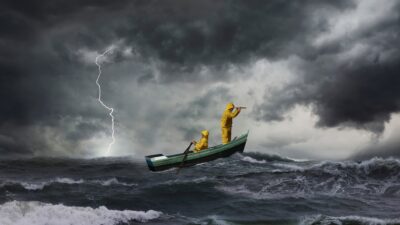 Imagem mostra duas pessoas, vestindo macacões amarelos, em um barquinho no meio de uma tempestade no mar. É possível ver ondas altas, nuvens escuras e relâmpagos. A pessoa à frente está com binóculos, olhando para a frente