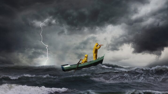 Imagem mostra duas pessoas, vestindo macacões amarelos, em um barquinho no meio de uma tempestade no mar. É possível ver ondas altas, nuvens escuras e relâmpagos. A pessoa à frente está com binóculos, olhando para a frente