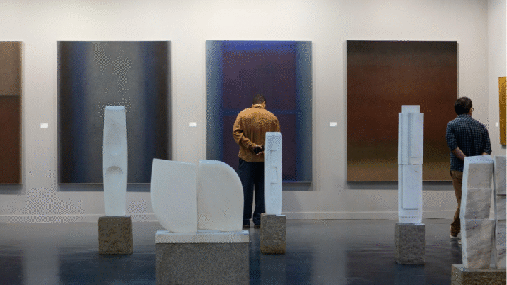 Homem de costas em frente a um de três quadros em um espaço expositivo. Em torno dele, há esculturas brancas.