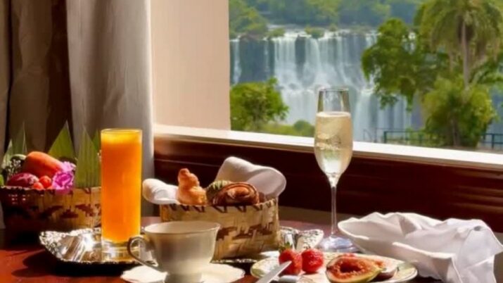 Mesa posta com brunch, suco, espumante e comida, à beira de uma janela; ao fundo, as Cataratas do Iguaçu