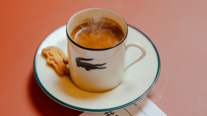 café expresso em xícara branca com logo de crocodilo da marca estampada no centro do objeto, sobre pires branco; ao lado, biscoitinho acompanha o café, também em formato de crocodilo