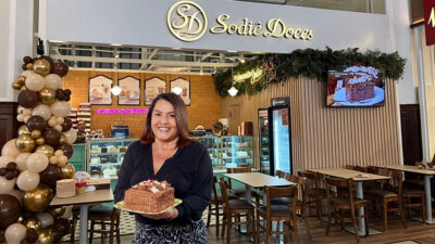 Montagem ilustrativa mostra Cleusa Maria segurando um bolo em primeiro plano, diante do interior de uma loja da Sodiê Doces. Ao fundo são vistos vitrines de doces, mesas, cadeiras, decoração com balões e a identidade visual da marca. A composição foi criada digitalmente e não corresponde a uma fotografia única de uma situação real.