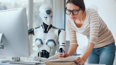 Pessoa trabalha ao lado de um robô humanoide em frente a um computador. A imagem ilustra os impactos da inteligência artificial na produtividade, na jornada de trabalho de 32 horas e nas discussões sobre o fim da escala 6x1.