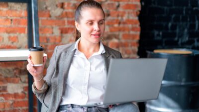 Mulher apreciando um café enquanto trabalha remotamente em um laptop dentro de casa.