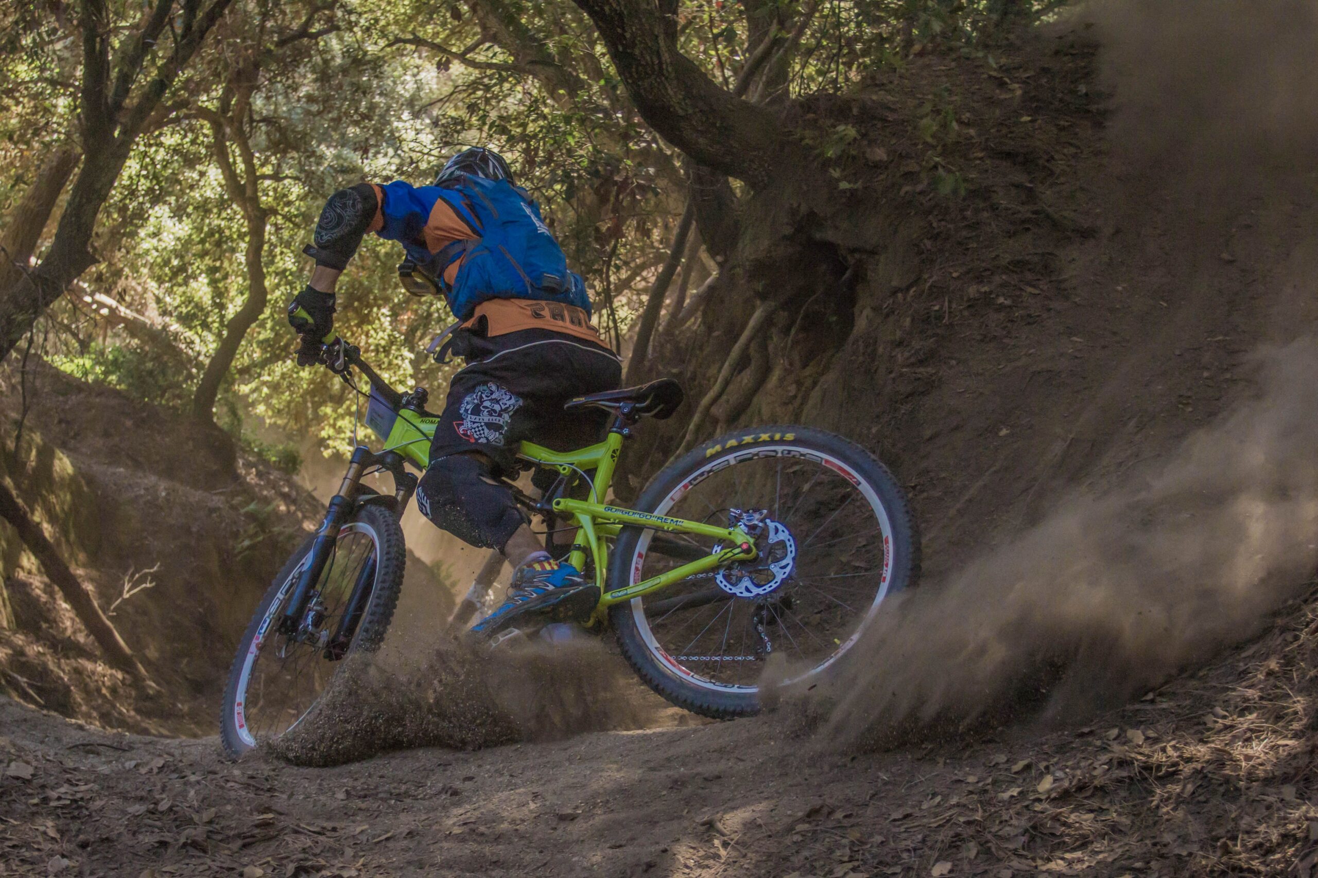 Ciclista de mountain bike faz curva em trilha de terra entre árvores, levantando poeira.