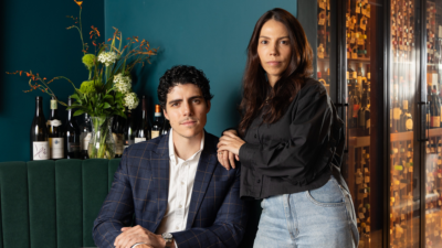 Retrato de Danyel Steinle e Tássia Magalhães, sócios do Lita, em São Paulo. Ele é um homem branco de cabelos escuros, que usa uma camisa branca e paletó e aparece sentado; apoiada em seu ombro, Tássia é uma mulher de pele parda clara e cabelos lisos e soltos, que usa uma blusa branca e uma calça jeans