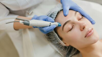 Paciente mulher branca deitada de olhos fechados com cabelos cobertos por uma touca. Mãos de um profissional se aproximam do rosto dela com um aparelho de estética facial