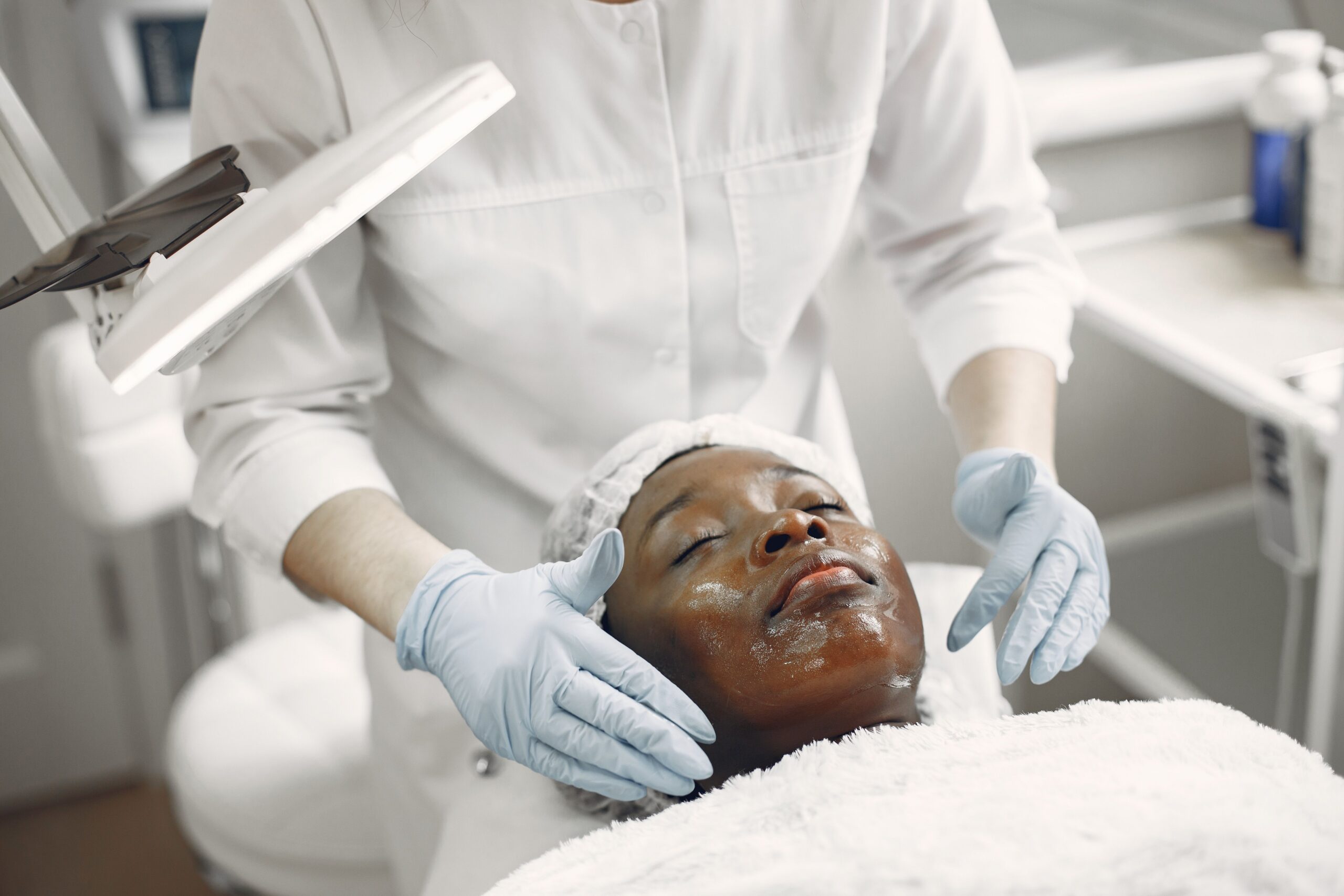 Paciente negra do sexo feminino aparece deitada em maca enquanto profissional de cor branca trata a pele de seu rosto