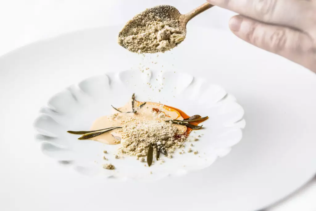 Mão polvilha pó sobre prato branco com preparo gourmet e ervas.