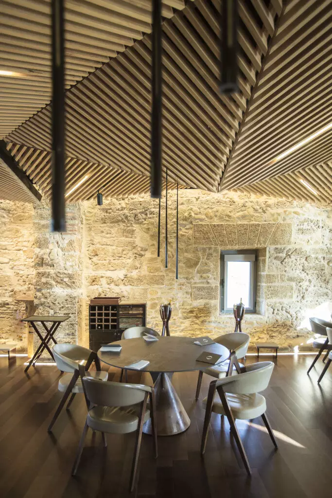 Sala de restaurante com mesa redonda, cadeiras ao redor, paredes de pedra e teto com ripas de madeira.