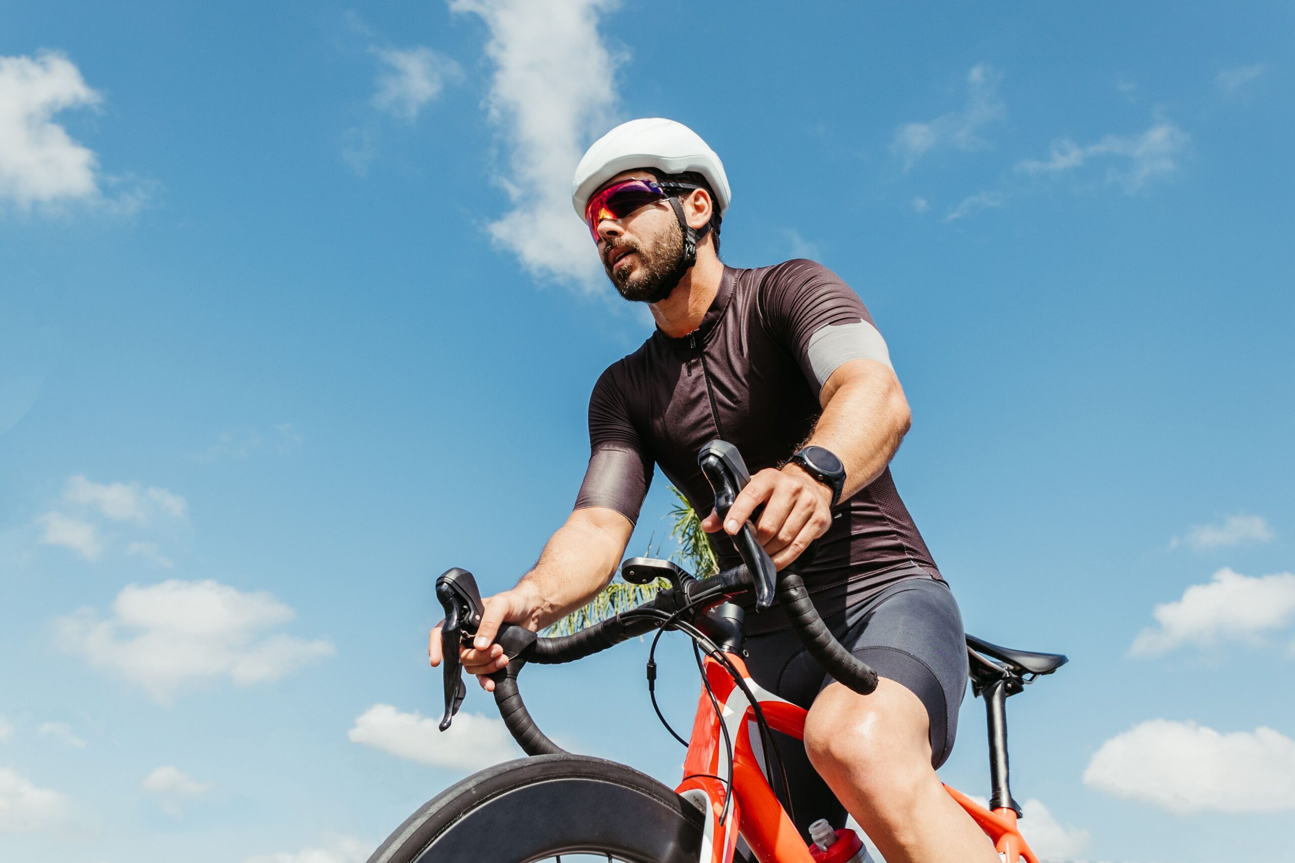 Homem de capacete e óculos escuros pedala uma bicicleta de estrada sob céu azul.