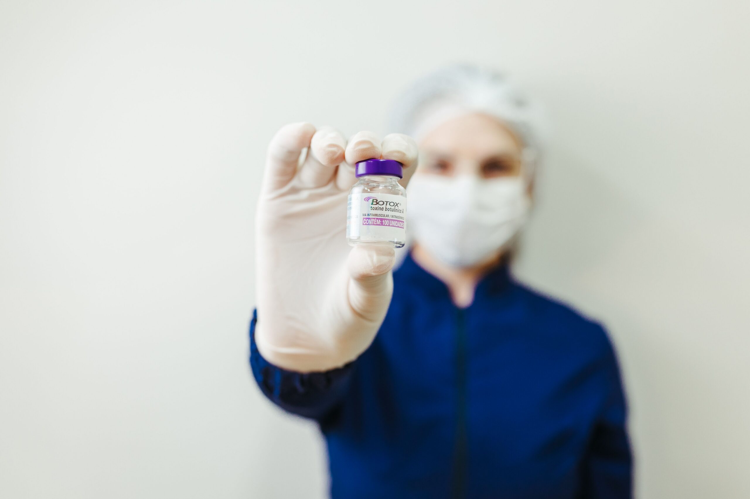 Pessoa com EPI, luvas, touca e máscara, aparece ao fundo, desfocada, segurando, à frente, um frasco de Botox