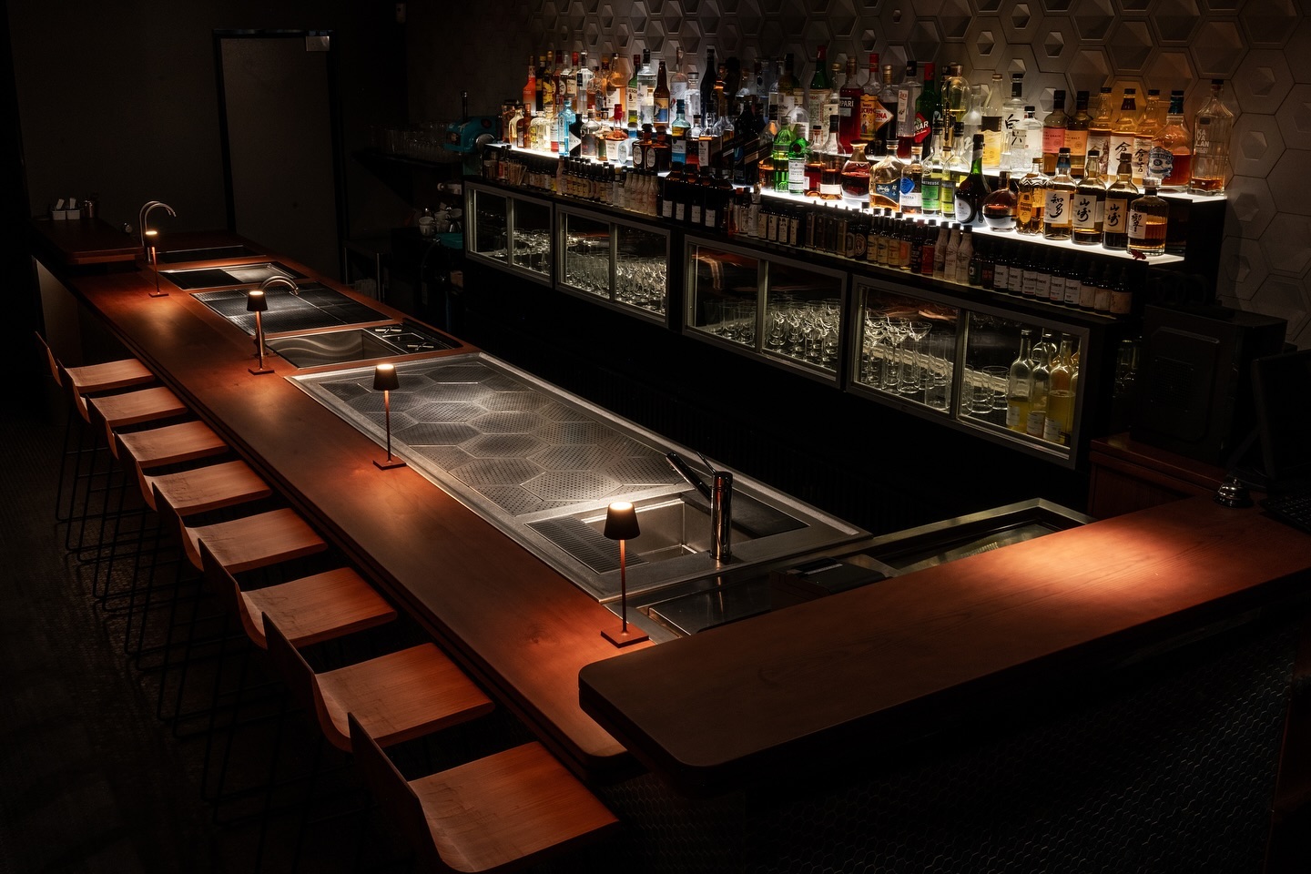 Balcão de bar em madeira com banquetas, pias embutidas e prateleiras cheias de garrafas ao fundo