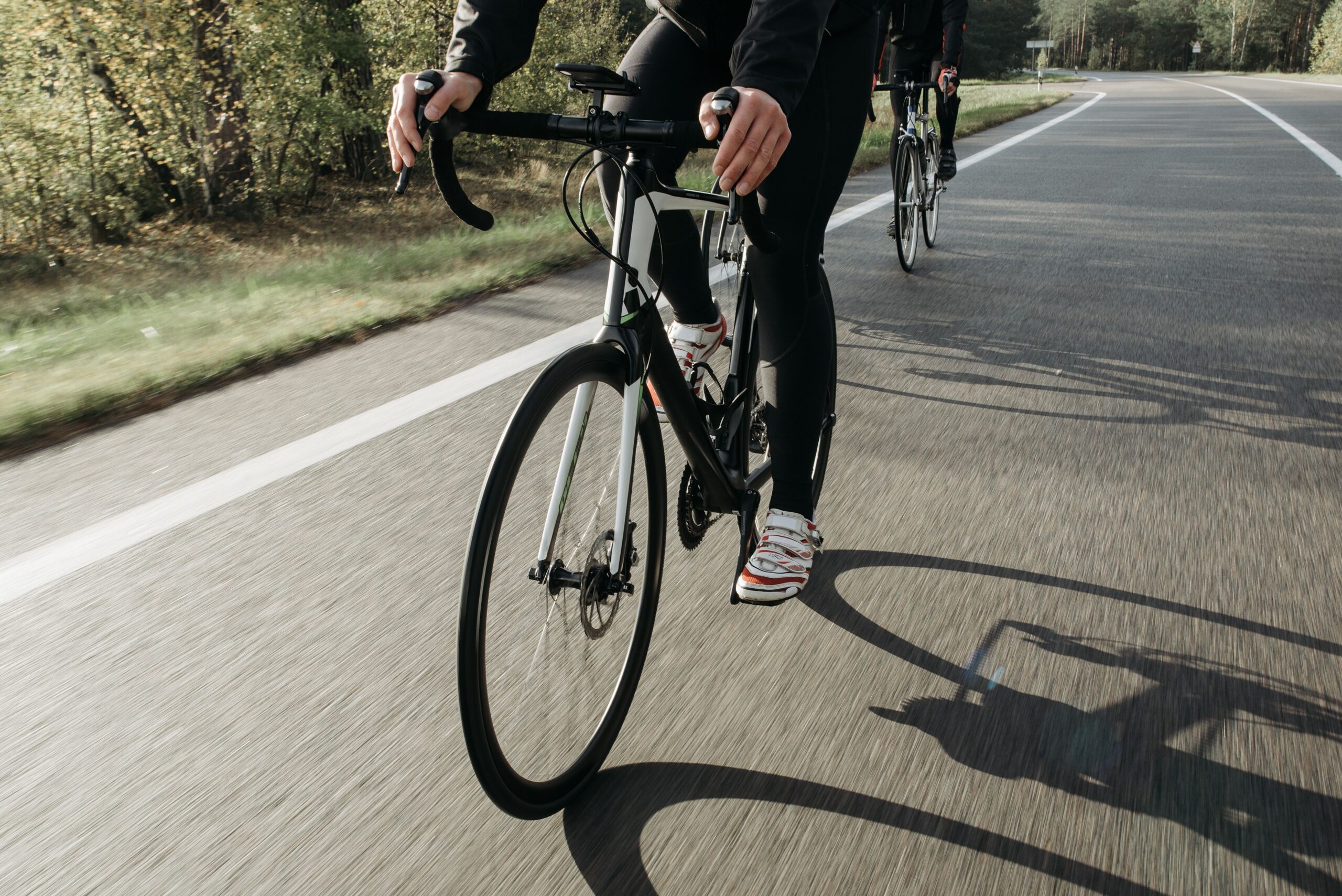 Dois ciclistas em bicicletas de estrada pedalando em uma via asfaltada cercada por árvores.