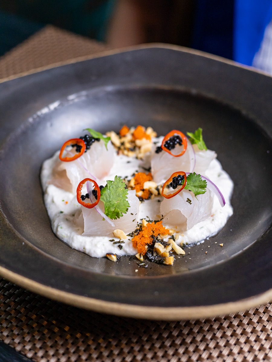 Prato fundo preto com pedaços de peixe cru sobre creme branco, decorados com pimenta, ervas e ovas.