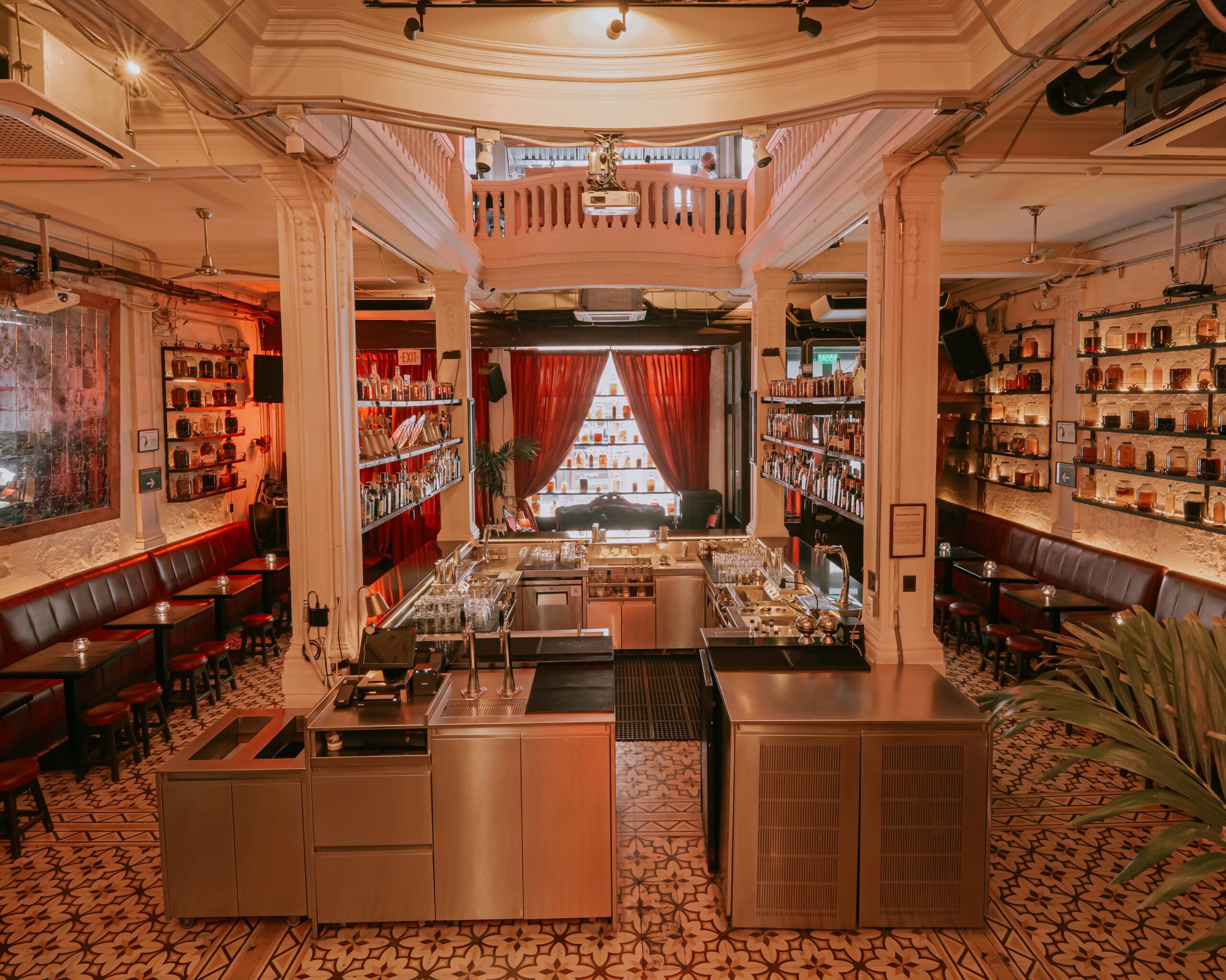 Interior de bar com balcão central, prateleiras de garrafas nas paredes e cortinas vermelhas ao fundo.