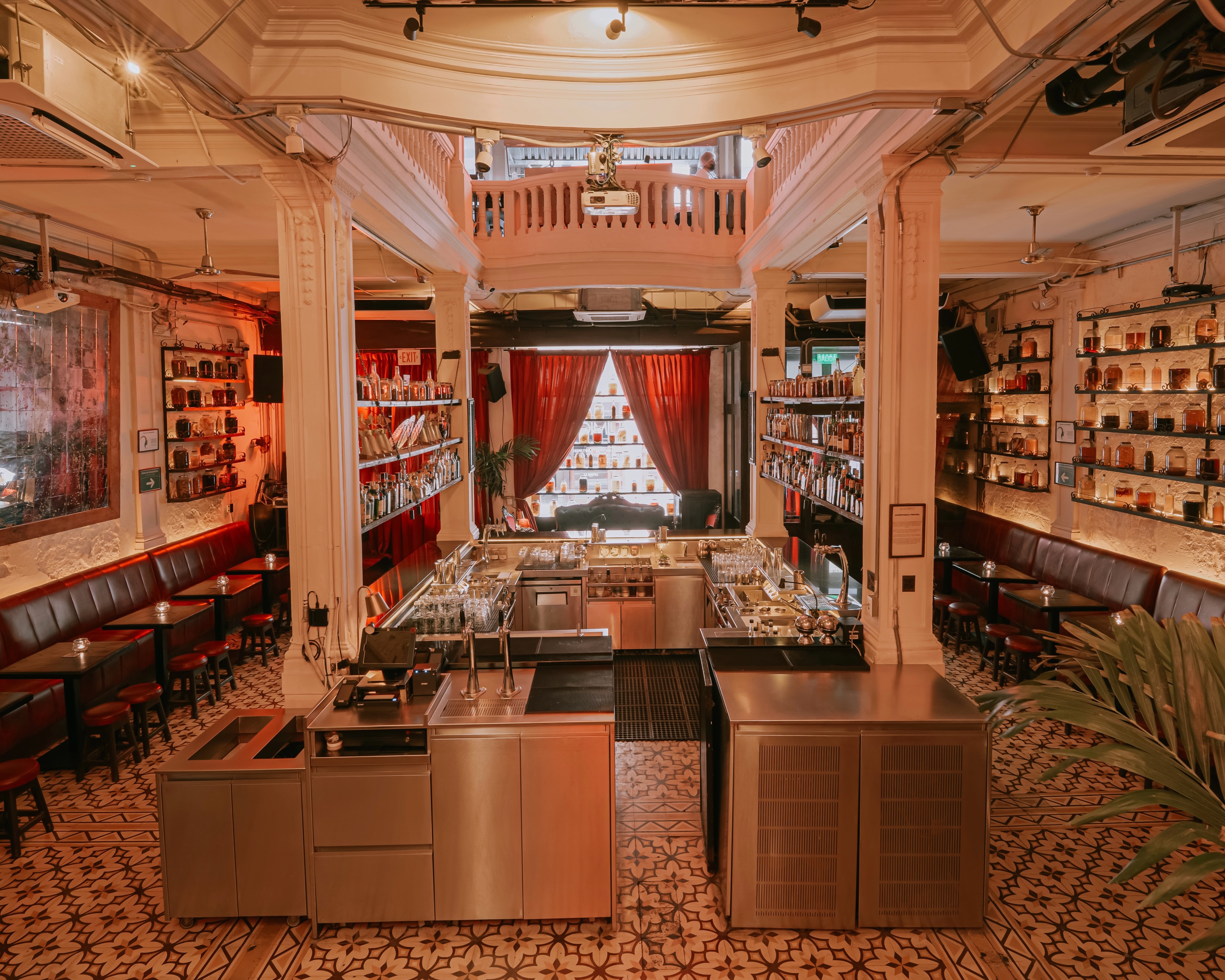Interior de bar com balcão central, prateleiras de garrafas nas paredes e cortinas vermelhas ao fundo.