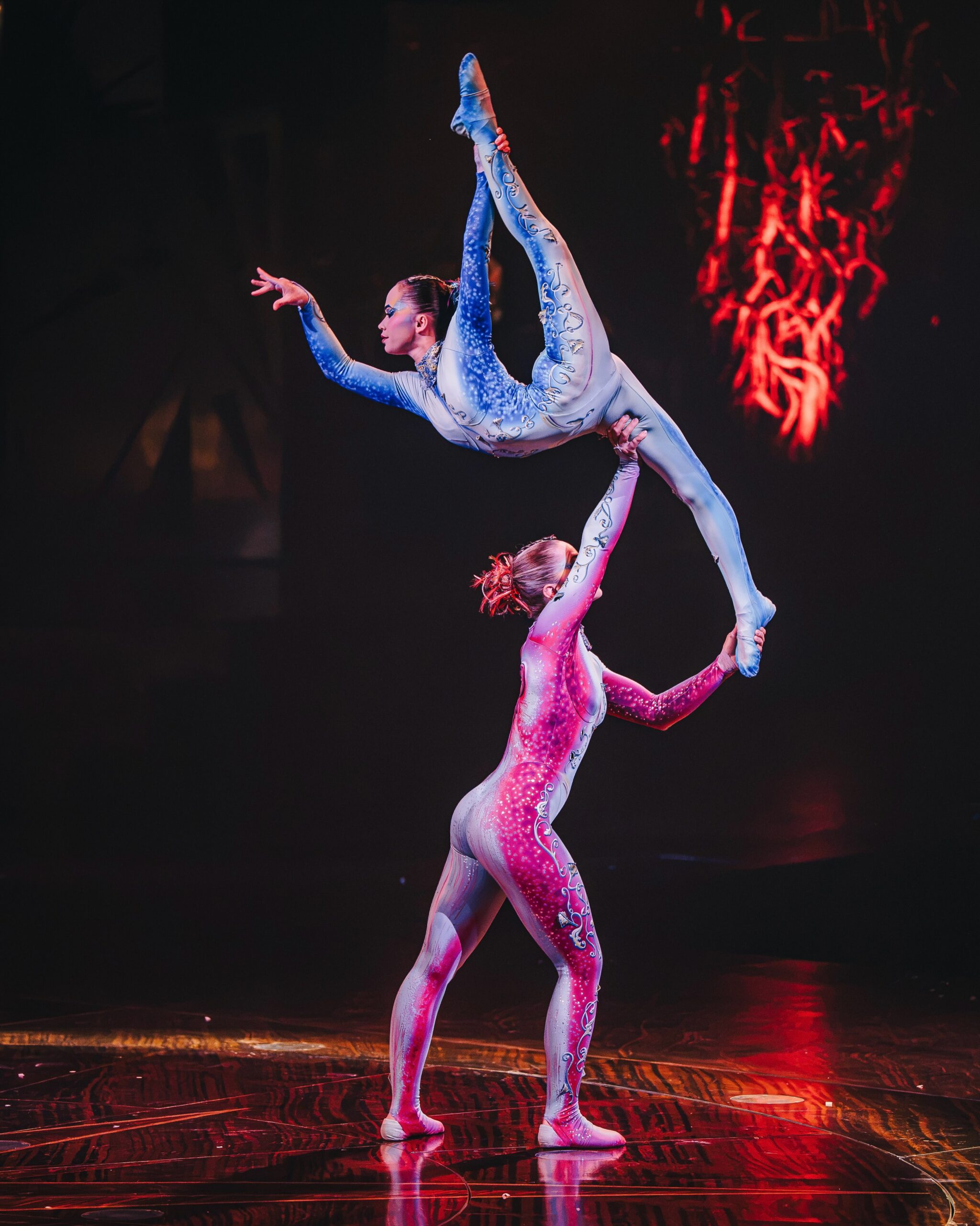 Duas acrobatas em figurinos coloridos fazem uma pose de equilíbrio em um palco escuro.