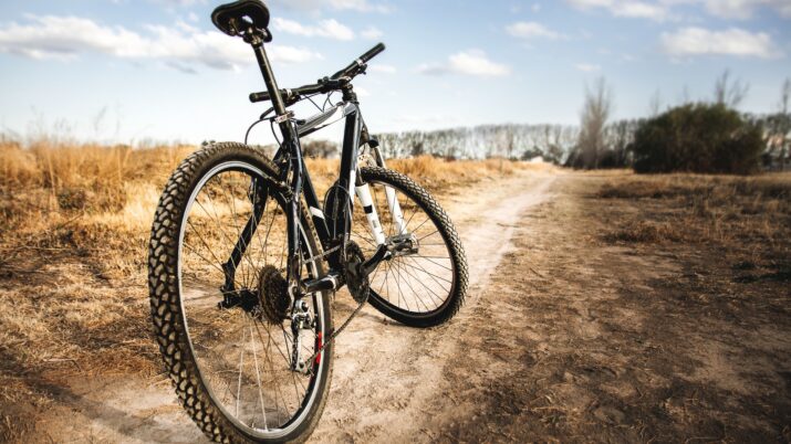 Bicicleta apoiada em uma trilha de terra em área aberta, com céu parcialmente nublado ao fundo.