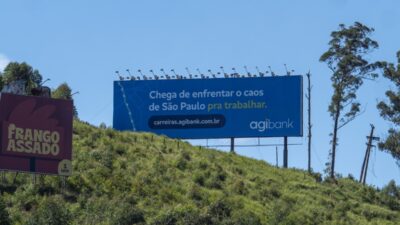 Dois outdoors à beira de uma rodovia, um com anúncio da Agibank e outro de frango assado, em uma encosta gramada.