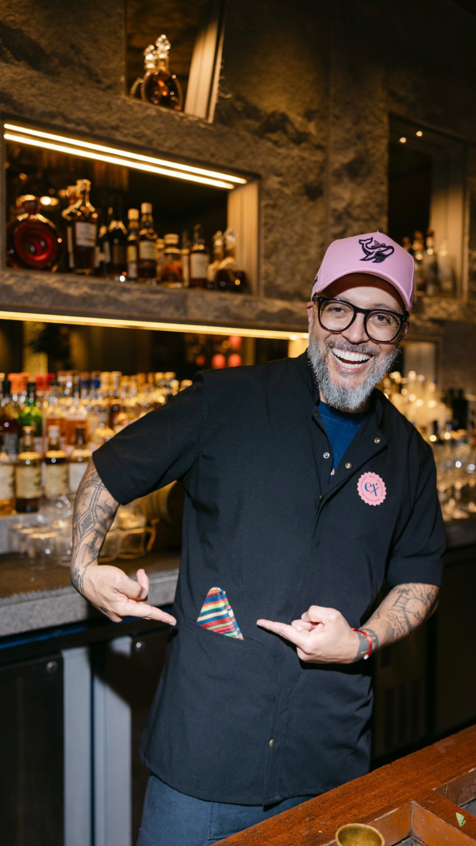 Homem sorridente de boné rosa e óculos, em frente a prateleiras com garrafas em um bar.