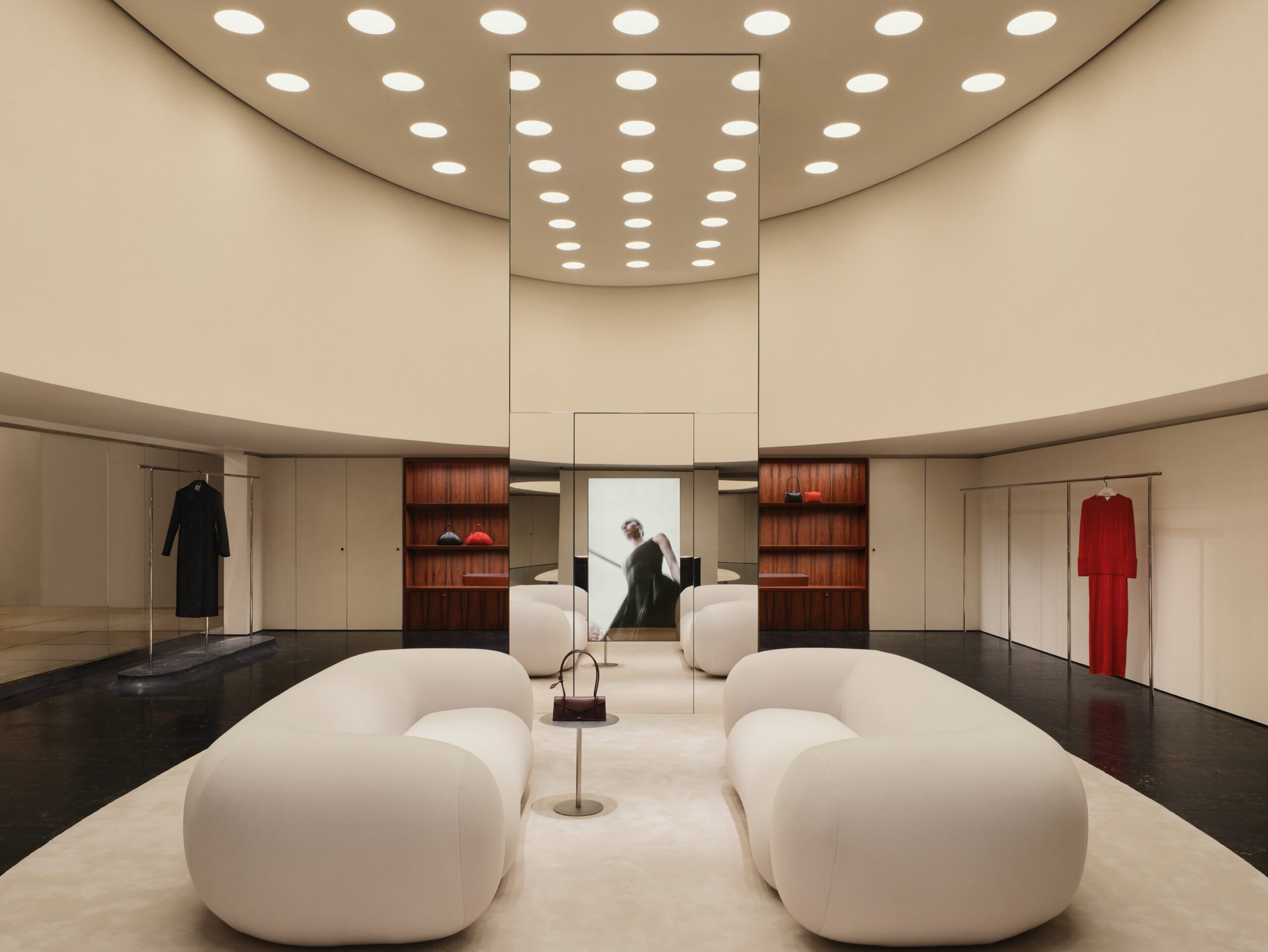 Interior de uma loja de luxo com espelhos, iluminação no teto, poltronas brancas, roupas expostas e bolsas em prateleiras.