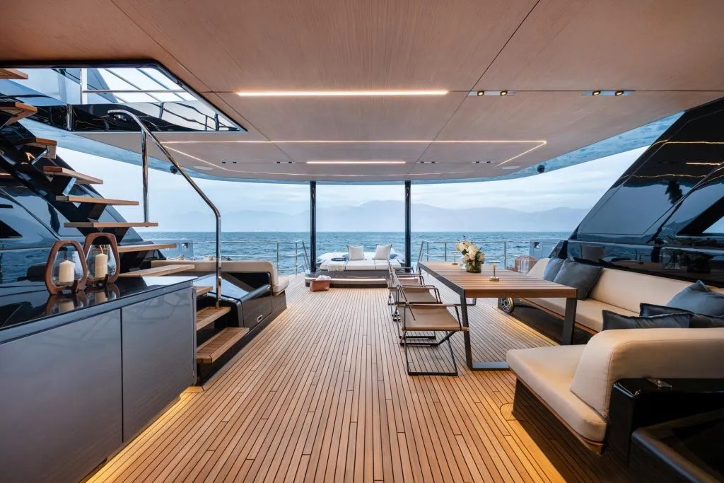 Convés de um iate de luxo com piso de madeira, sofás, mesa e vista aberta para o mar.