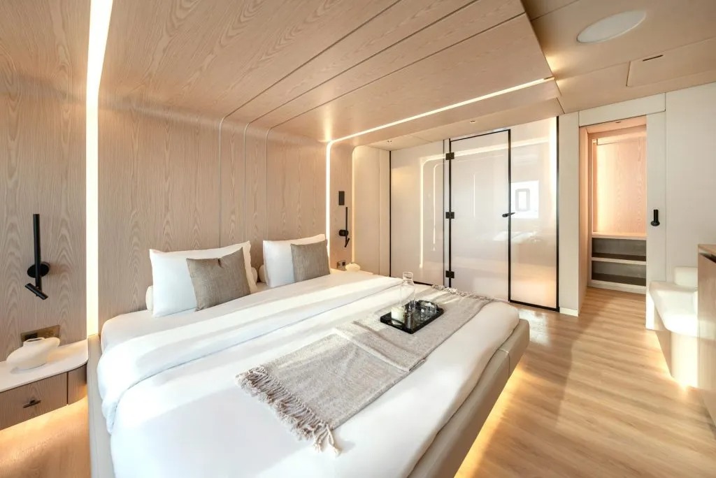 Quarto moderno com cama grande, paredes e teto em madeira clara, iluminação embutida e porta de vidro ao fundo.