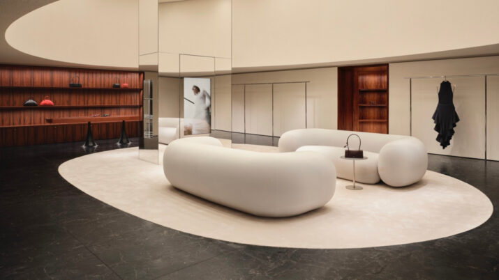 Interior de loja com sofá branco curvo, piso escuro, paredes espelhadas e bolsas e roupas expostas nas prateleiras.