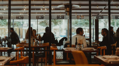 Interior de restaurante com mesas postas e clientes sentados conversando ao fundo, vistos através de divisórias de vidro.