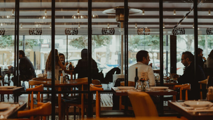 Interior de restaurante com mesas postas e clientes sentados conversando ao fundo, vistos através de divisórias de vidro.
