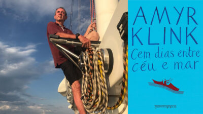 Homem sentado na estrutura de um veleiro, com cordas enroladas ao lado, sob céu azul com nuvens.