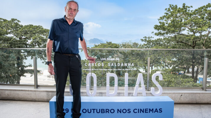 Homem em pé ao lado de letreiro com o nome O Dia, em uma varanda com vista para árvores e céu azul.