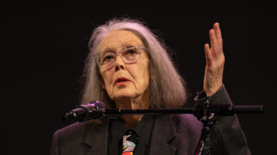 Anne Carson de óculos fala ao microfone com a mão levantada, em fundo escuro.