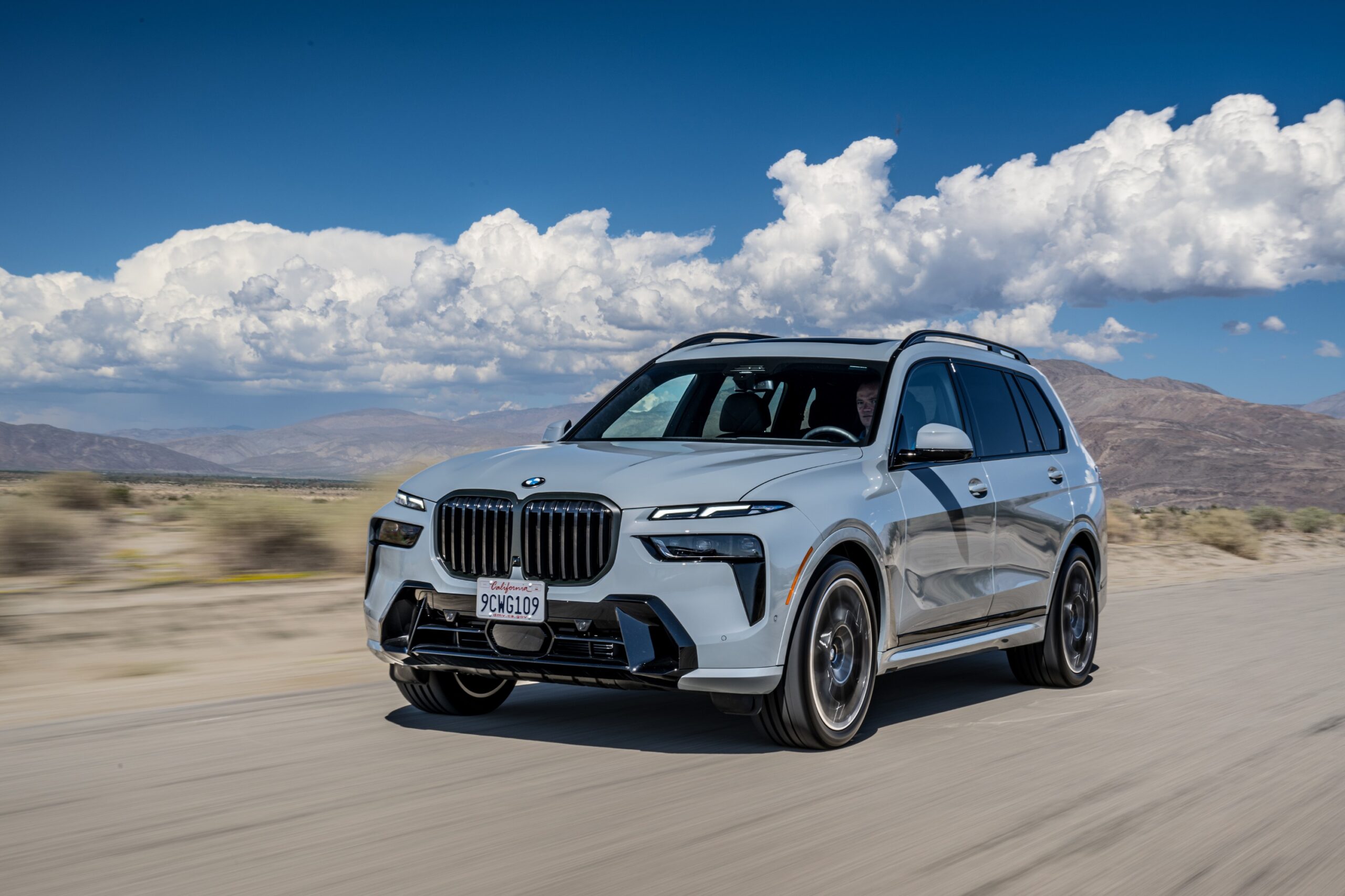 SUV BMW branco em movimento em uma estrada, com montanhas e céu azul com nuvens ao fundo.