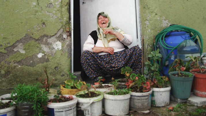 Mulher sentada na entrada de uma casa, cercada por vasos de plantas e um muro verde descascado.