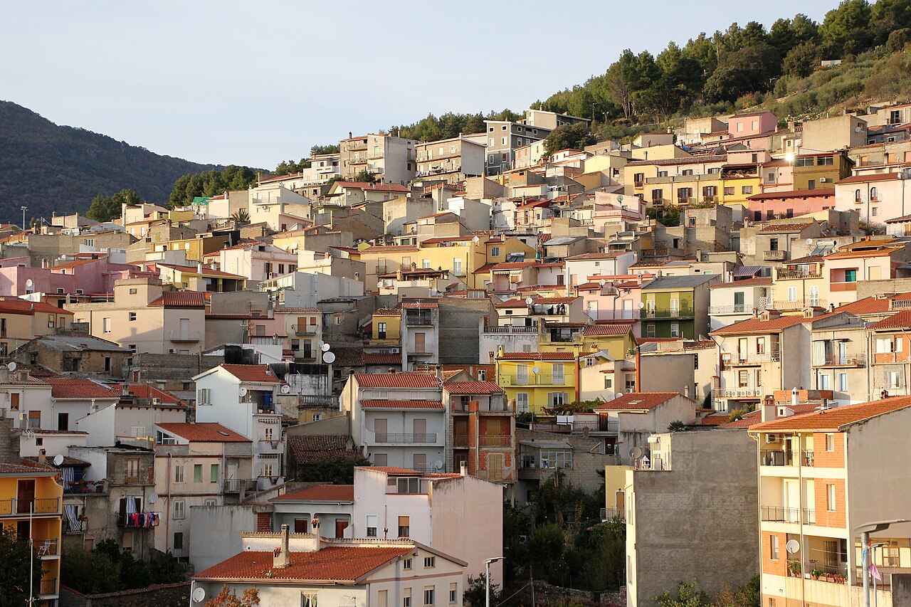Casas coloridas e prédios baixos espalhados por uma encosta, com árvores e montanhas ao fundo.