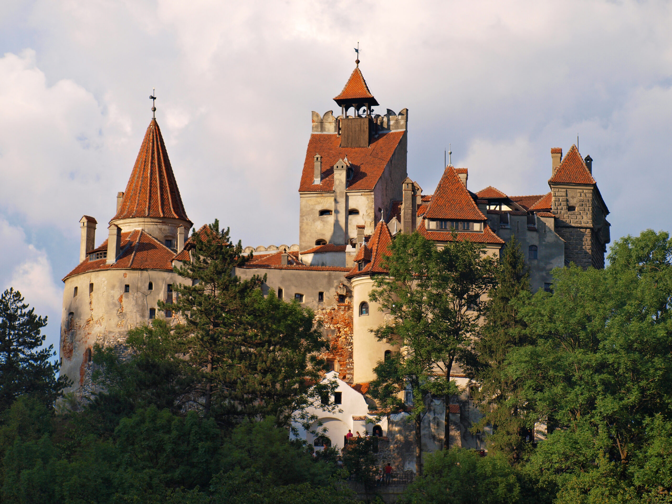 Castelo medieval com torres e telhados vermelhos cercado por árvores sob céu nublado