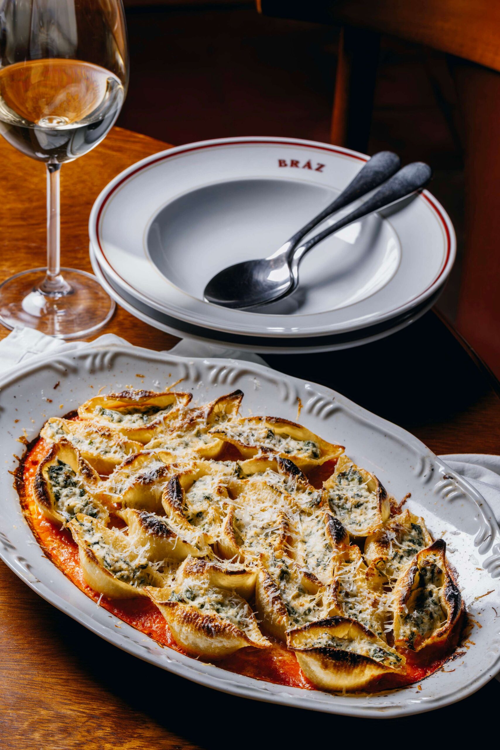 Prato com massa gratinada, taça de vinho e pratos empilhados sobre mesa de madeira.