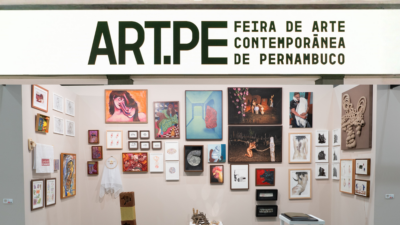 Estande da Art.PE com várias obras emolduradas e peças tridimensionais expostas em uma parede branca.