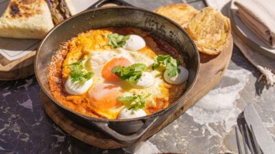 Frigideira com ovos sobre molho vermelho, com folhas de coentro e pães tostados ao lado.