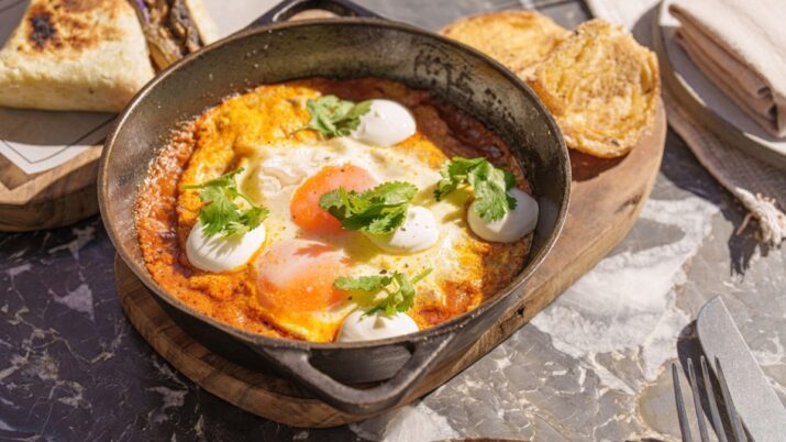 Frigideira com ovos sobre molho vermelho, com folhas de coentro e pães tostados ao lado.
