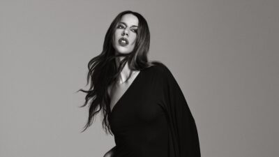 Mulher de cabelo longo e vestido escuro posa em fundo neutro, em preto e branco.