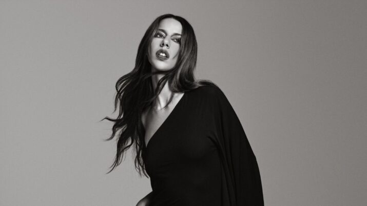 Mulher de cabelo longo e vestido escuro posa em fundo neutro, em preto e branco.