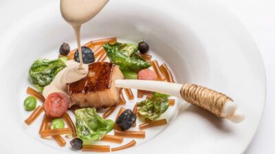 Prato branco com pedaço de peixe grelhado, legumes e molho sendo despejado por cima.