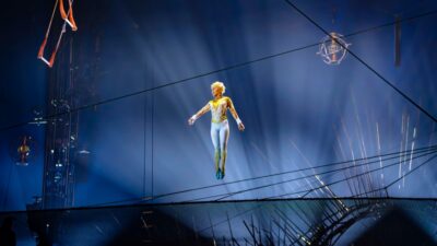 Artista com figurino branco e dourado aparece suspensa no ar sob luzes azuis em um palco de circo.